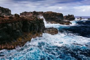 Waves crashing against rugged rocky cliffs under a cloudy sky.
