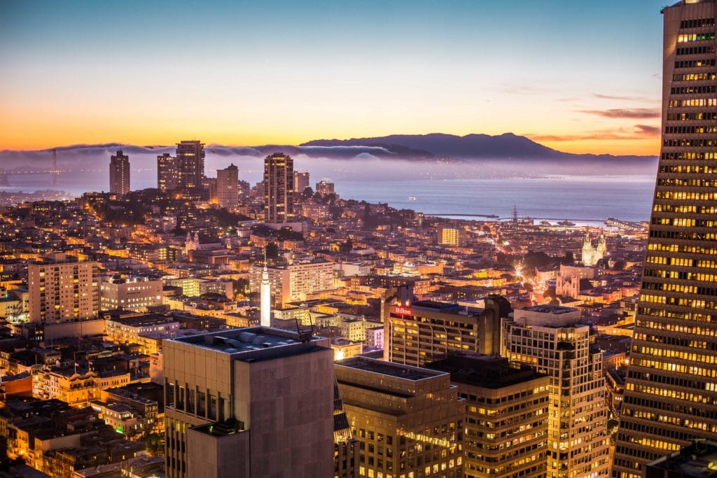 City skyline at sunset with illuminated buildings and distant mountains.