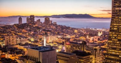 City skyline at sunset with illuminated buildings and distant mountains.