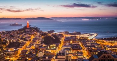 Cityscape at sunset with illuminated streets and a distant lighthouse.