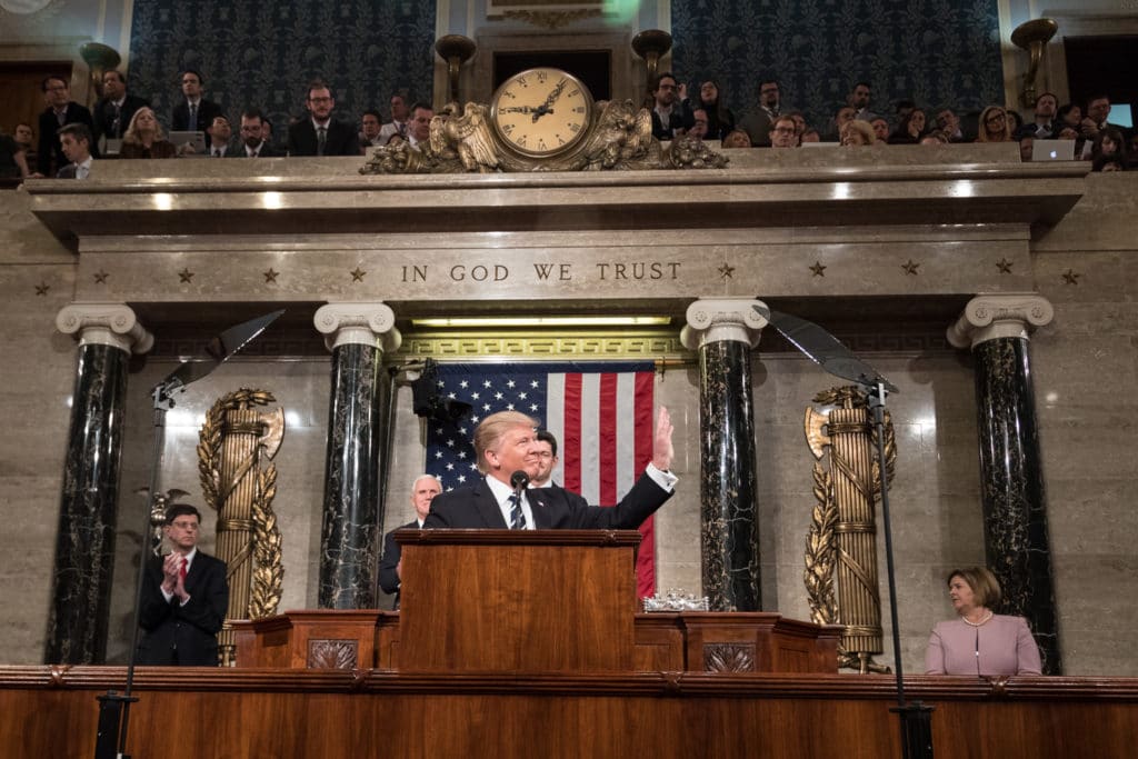 A man speaking at a podium in a formal government setting with an American flag behind.