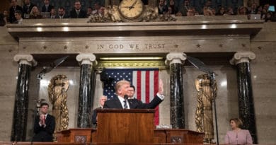 A man speaking at a podium in a formal government setting with an American flag behind.