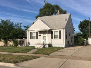 Small white house with a front porch and manicured lawn under blue sky.