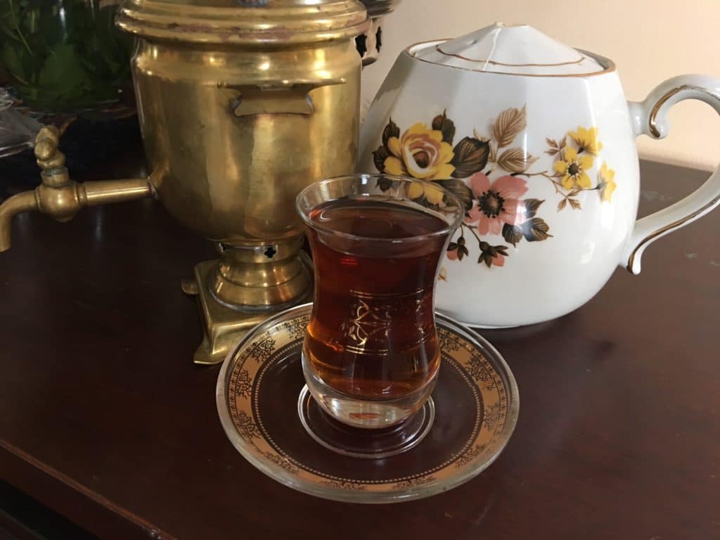 A glass of tea on a saucer with a floral teapot and brass samovar.