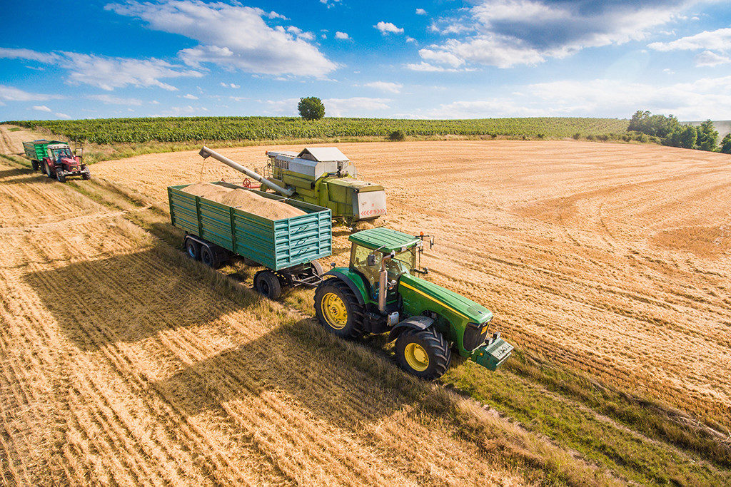 A tractor with a trailer harvesting crops in a vast field under a clear sky.