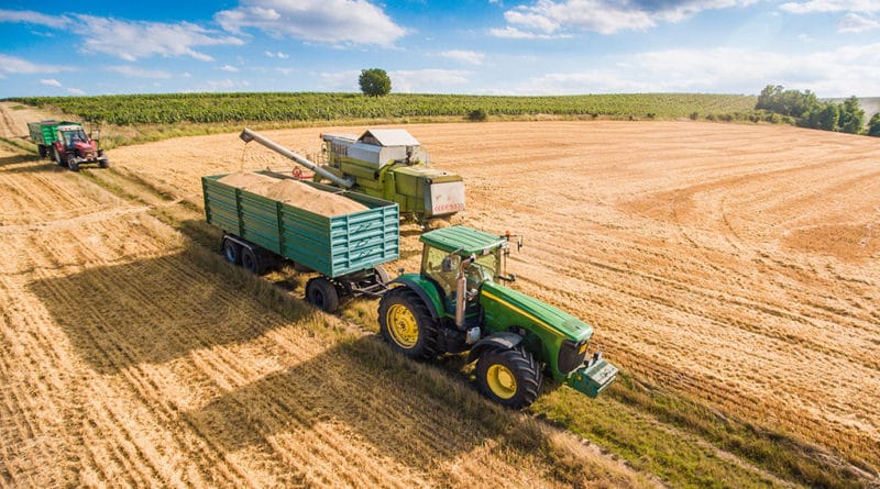 A tractor with a trailer harvesting crops in a vast field under a clear sky.