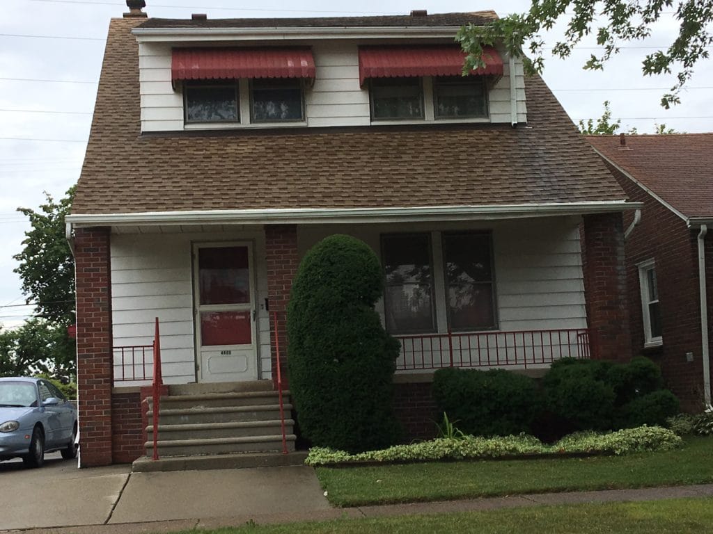 A modest brick house with a red awning and a trimmed shrub in front.