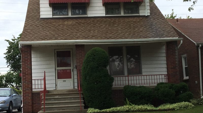 A modest brick house with a red awning and a trimmed shrub in front.