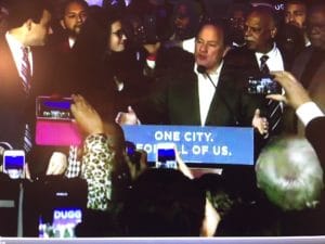 A man speaks at a podium with a sign that reads 'One City. One of Us.'