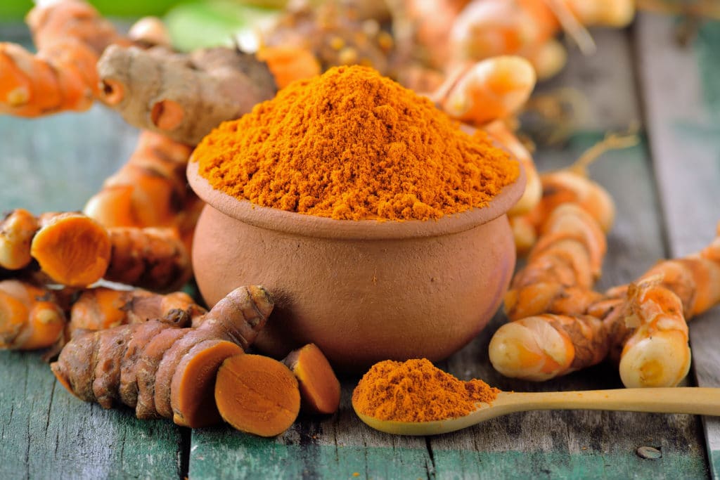 A bowl filled with bright orange turmeric powder with fresh turmeric roots around it.