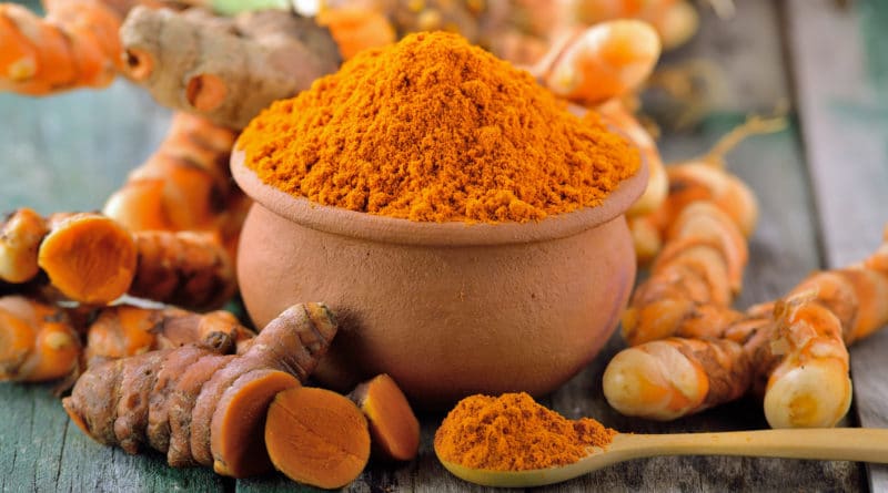 A bowl filled with bright orange turmeric powder with fresh turmeric roots around it.