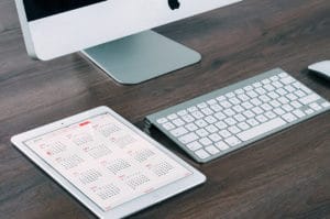 Tablet displaying a calendar next to a keyboard and monitor on a wooden desk.