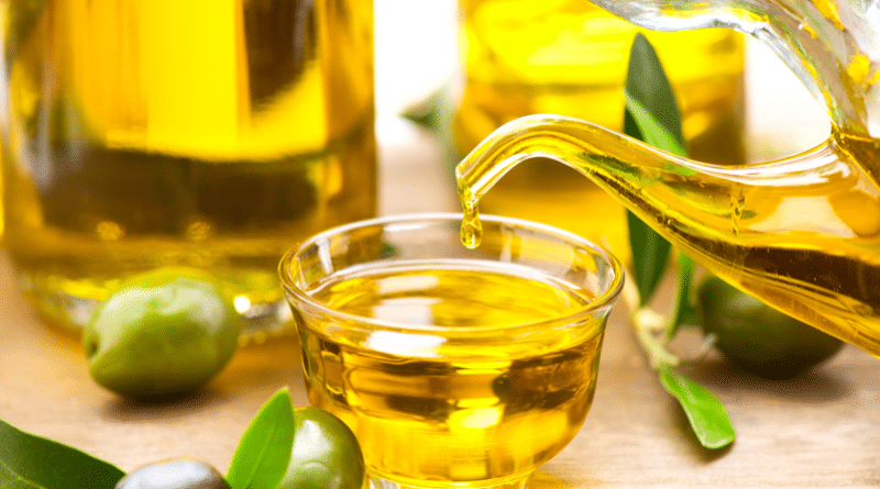 A small glass bowl filled with olive oil surrounded by green olives and olive branches.