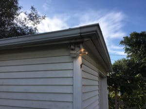 White exterior corner of a house with outdoor light and trees under a blue sky.