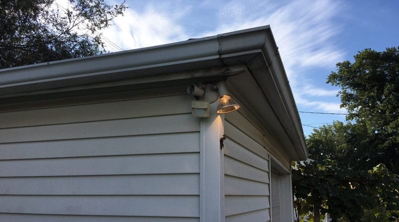 White exterior corner of a house with outdoor light and trees under a blue sky.