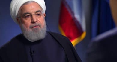 Portrait of a man in traditional attire with an Iranian flag in the background.