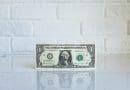 A one-dollar bill standing upright on a reflective surface against a white brick wall.