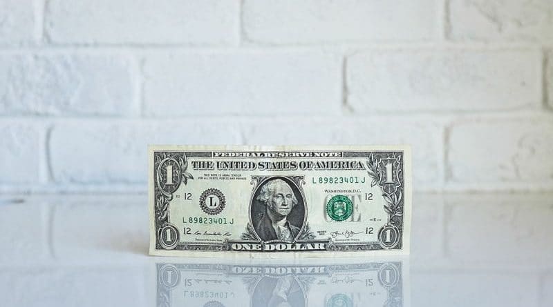 A one-dollar bill standing upright on a reflective surface against a white brick wall.