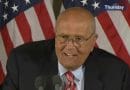 An elderly man speaking at a podium with American flags behind him.
