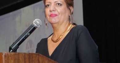 A woman speaking at a podium with a microphone, wearing a black dress and gold necklace.