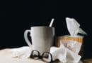 A coffee mug, glasses, and tissues on a table against a dark background.