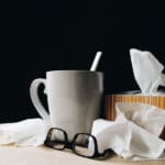 A coffee mug, glasses, and tissues on a table against a dark background.