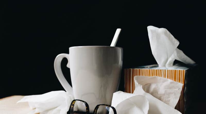 A coffee mug, glasses, and tissues on a table against a dark background.