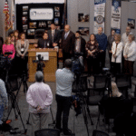 A speaker addresses an audience at a podium with cameras recording the event.