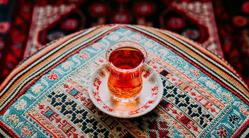 A glass of tea on a saucer sits on a patterned tablecloth.