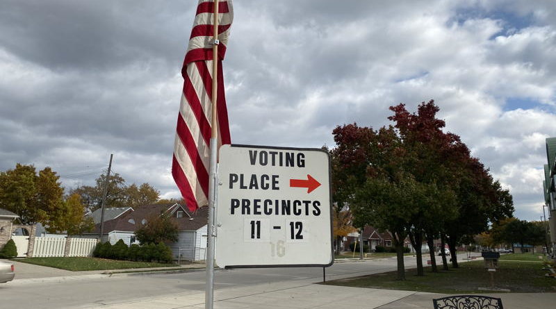 Sign for voting place and precincts with American flag and cloudy sky.