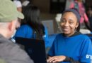 A young woman in a blue shirt smiles while using a laptop.