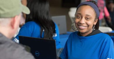 A young woman in a blue shirt smiles while using a laptop.