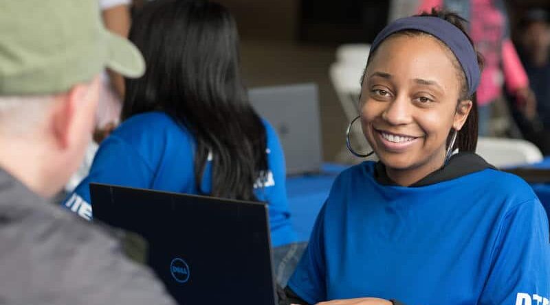 A young woman in a blue shirt smiles while using a laptop.