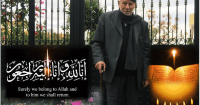 Elderly man in black coat and hat standing beside Islamic calligraphy outdoors.