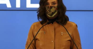 Person in a brown jacket speaking at a podium wearing a patterned face mask.