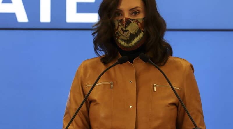 Person in a brown jacket speaking at a podium wearing a patterned face mask.