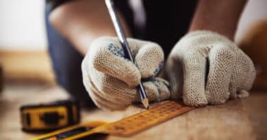 A person marking wood with a pencil and ruler while wearing gloves.