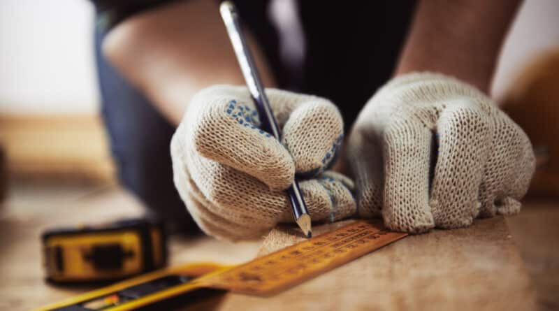 A person marking wood with a pencil and ruler while wearing gloves.
